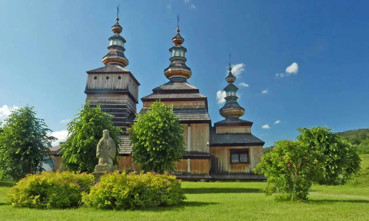 Drewniana cerkiew z trzema kopułami na tle błękitnego nieba, otoczona zielenią. Piękny widok w Beskidzie Niskim.