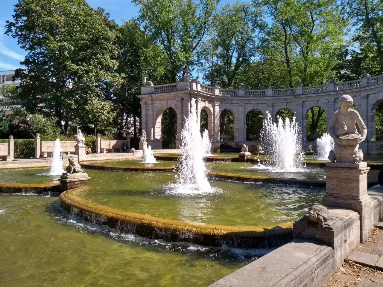 Wasserspiele im Volkspark Friedrichshain mit einer steinernen Bogenarchitektur im Hintergrund und einer Statue einer Frau mit einem Korb.