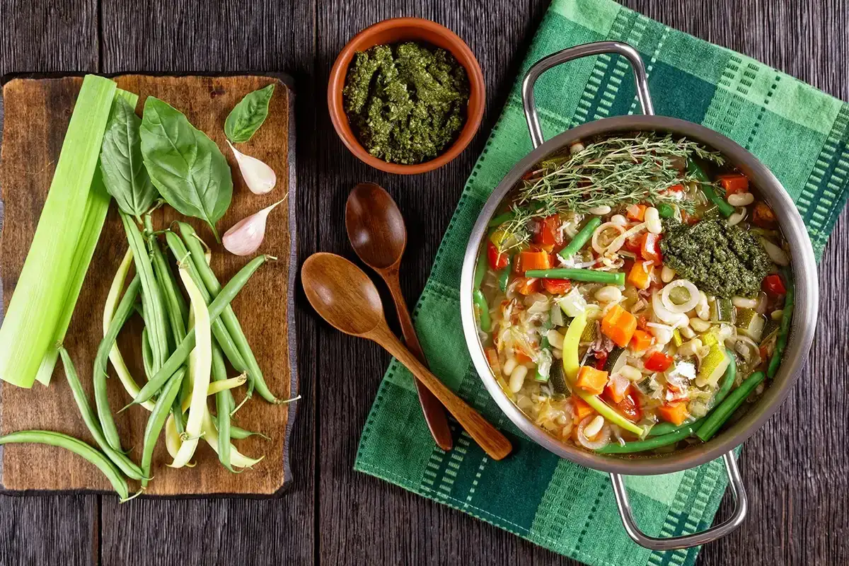 Une marmite de soupe au pistou d&eacute;bordante de l&eacute;gumes frais, avec une cuill&egrave;re de pesto vert vif. Des haricots verts, du c&eacute;leri et de l'ail sont dispos&eacute;s sur une planche en bois.