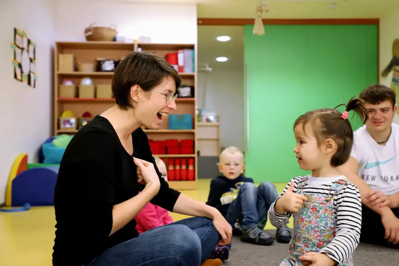 Eine Frau lacht und gestikuliert, w&auml;hrend sie mit einem Kleinkind sprechen &uuml;bt. Ein Junge und ein Mann sitzen im Hintergrund.