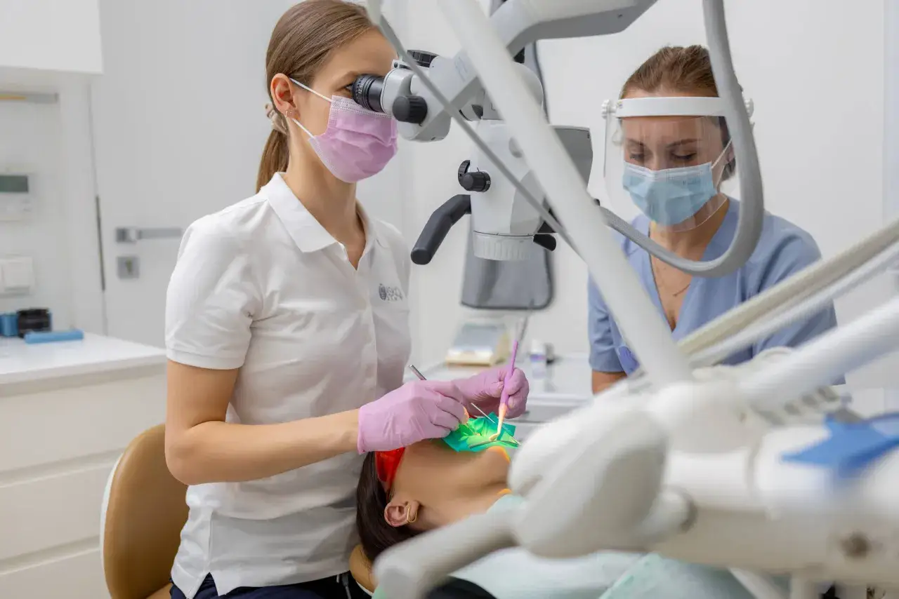 Dentystka w masce i rękawiczkach pracuje pod mikroskopem przy pacjencie.