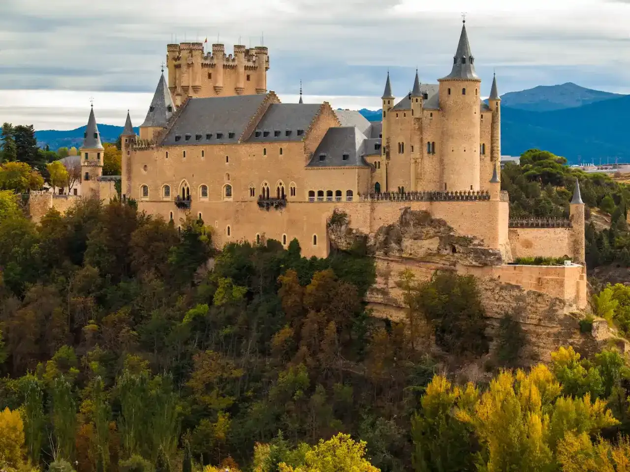 Majestuosos castillos medievales se alzan sobre un bosque otoñal, con torres y murallas de piedra dorada bajo un cielo nublado.