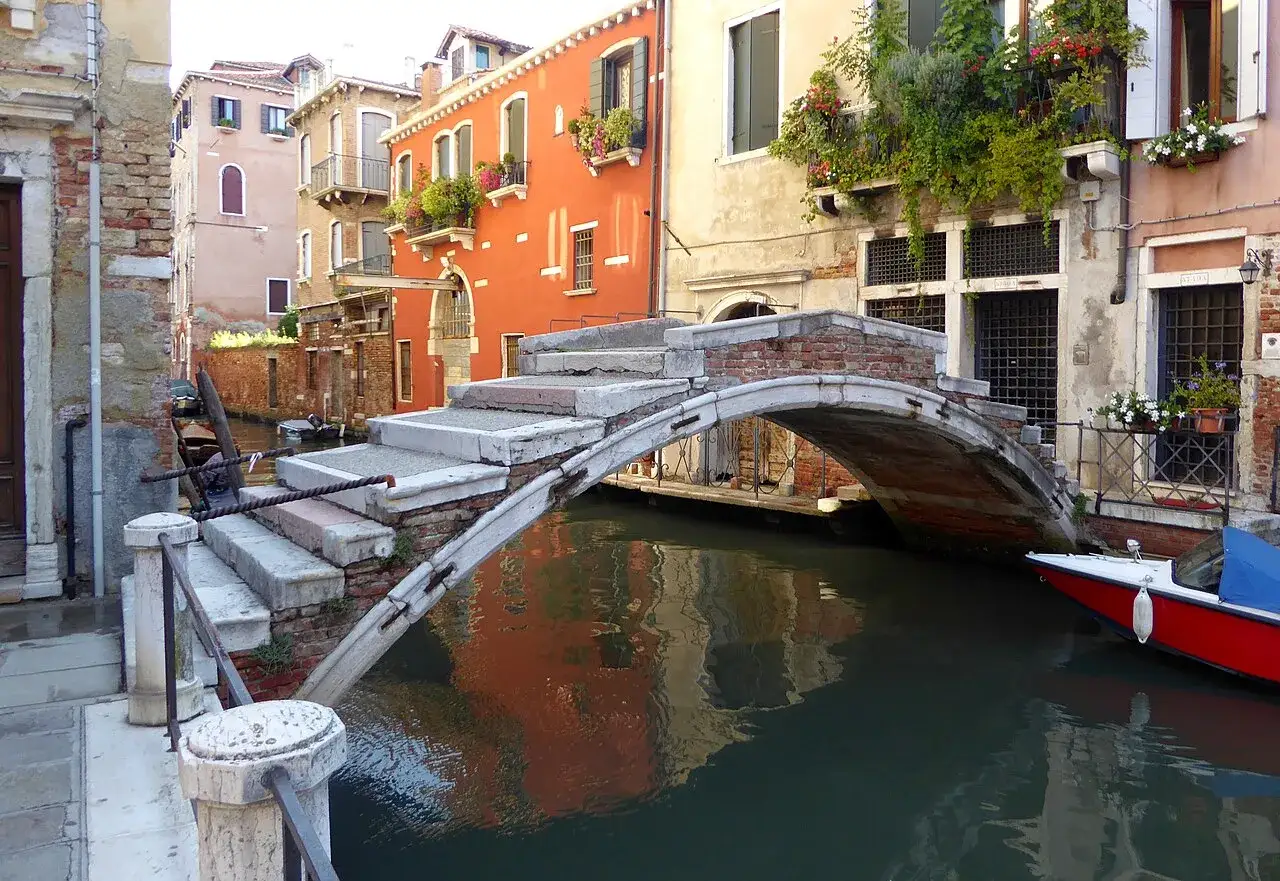 Un antico ponte a Venezia senza parapetto, con gradini in pietra e mattoni, riflette i colori degli edifici circostanti nell'acqua scura del canale.
