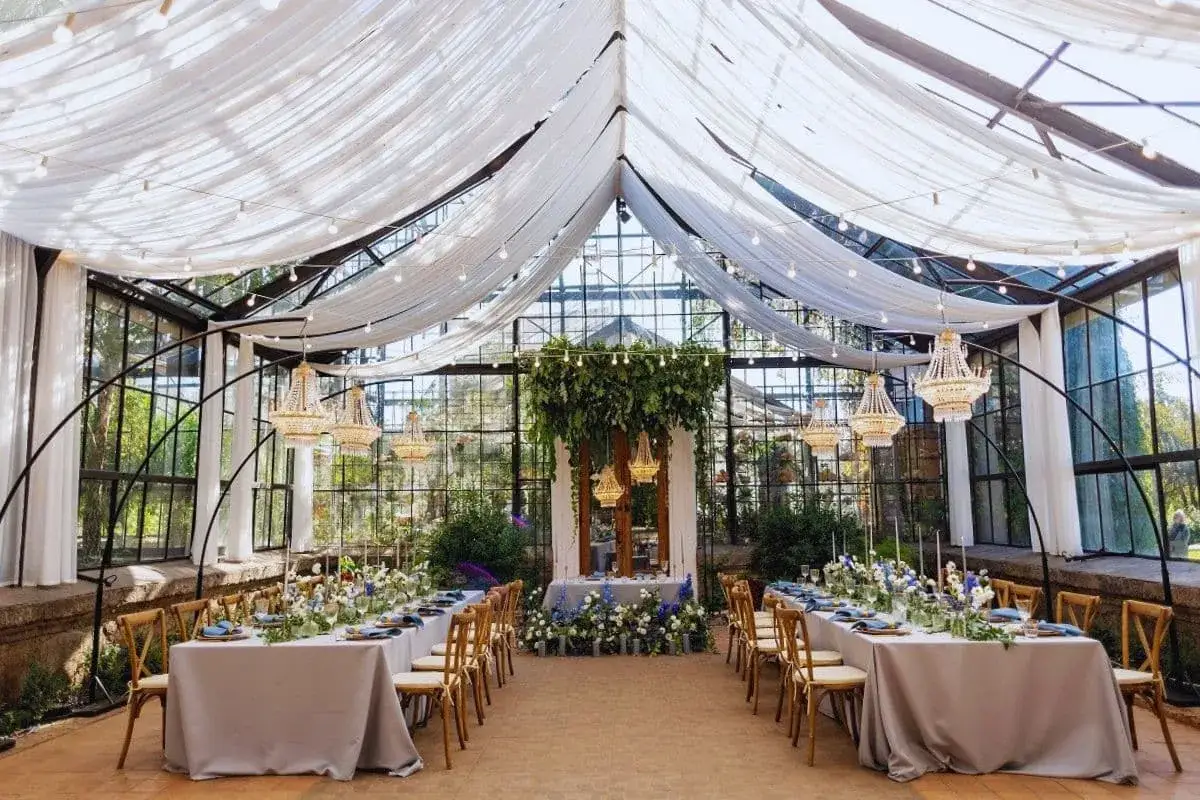 Magnifique serre aménagée pour un mariage de 50 personnes, avec des tables dressées, des lustres étincelants et une décoration florale luxuriante.