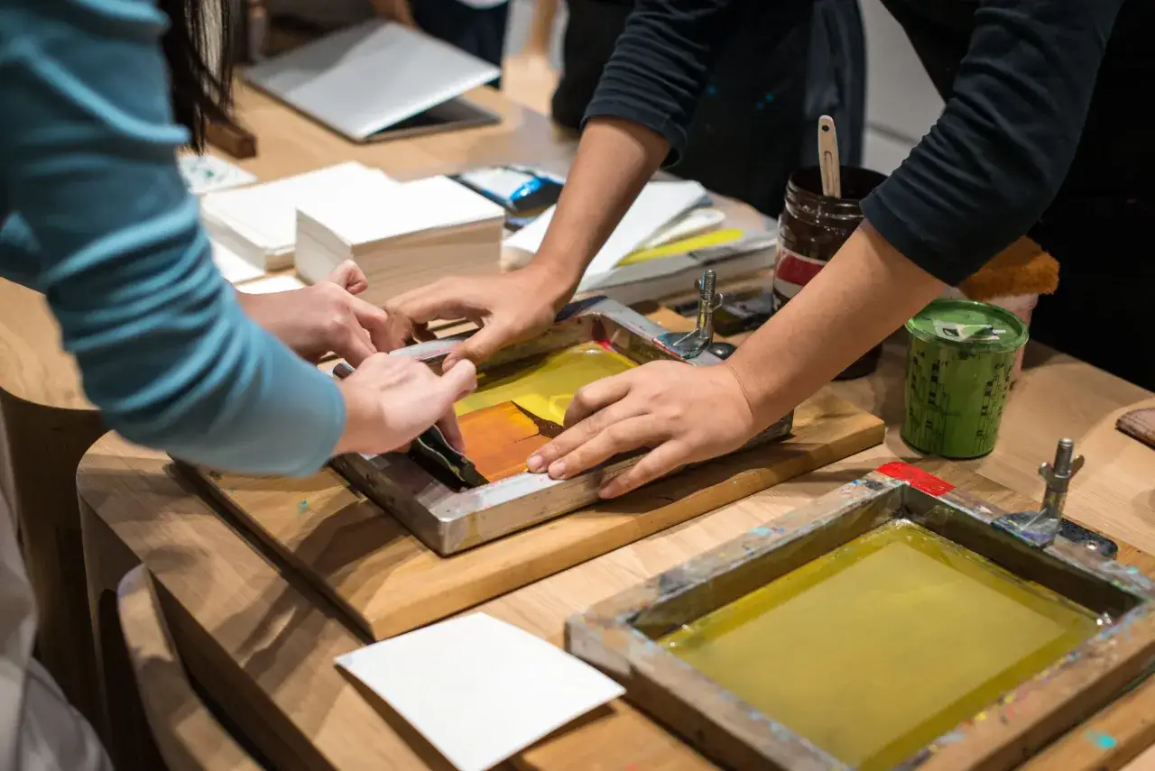 Des mains travaillent sur un &eacute;cran de s&eacute;rigraphie, pr&eacute;parant une **formation design textile**. Des pots de peinture et du papier sont visibles.