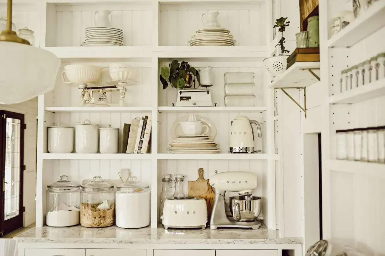 A bright, white kitchen with open shelving showcasing vintage scales, ceramic canisters, books, and cream-colored appliances like a Smeg mixer and toaster, perfect for an affordable kitchen makeover.