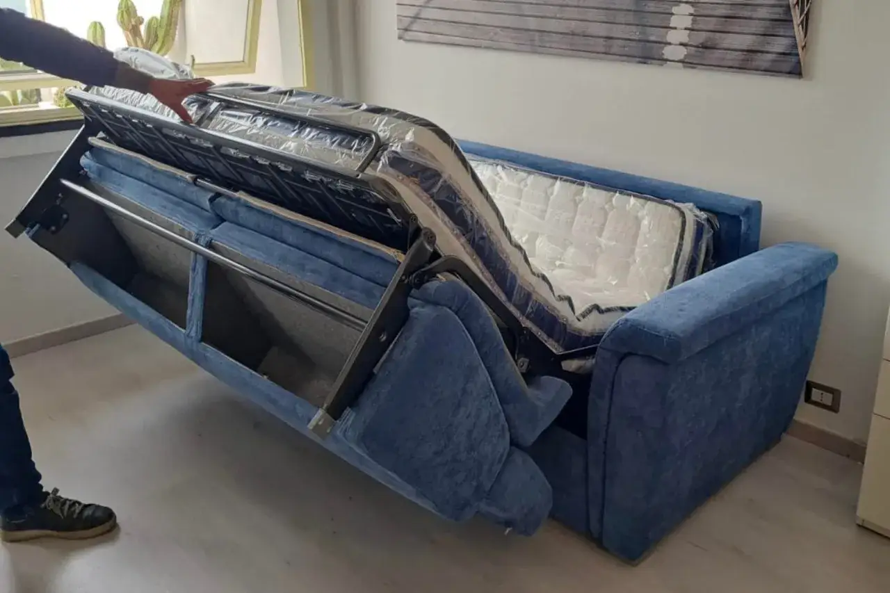 Un braccio solleva il meccanismo di un divano letto blu, mostrando la struttura metallica e il materasso. Riparazione meccanismo divano letto in corso.