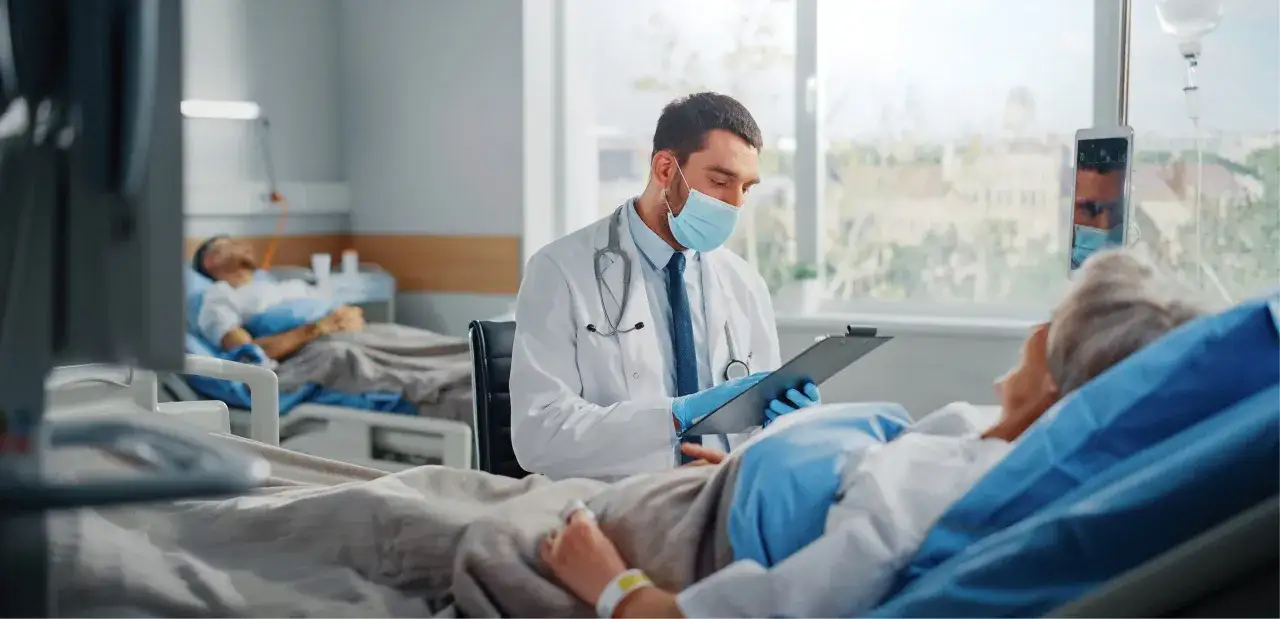 Lekarz w masce i rękawiczkach rozmawia z pacjentką podczas hospitalizacji. W tle inni pacjenci.