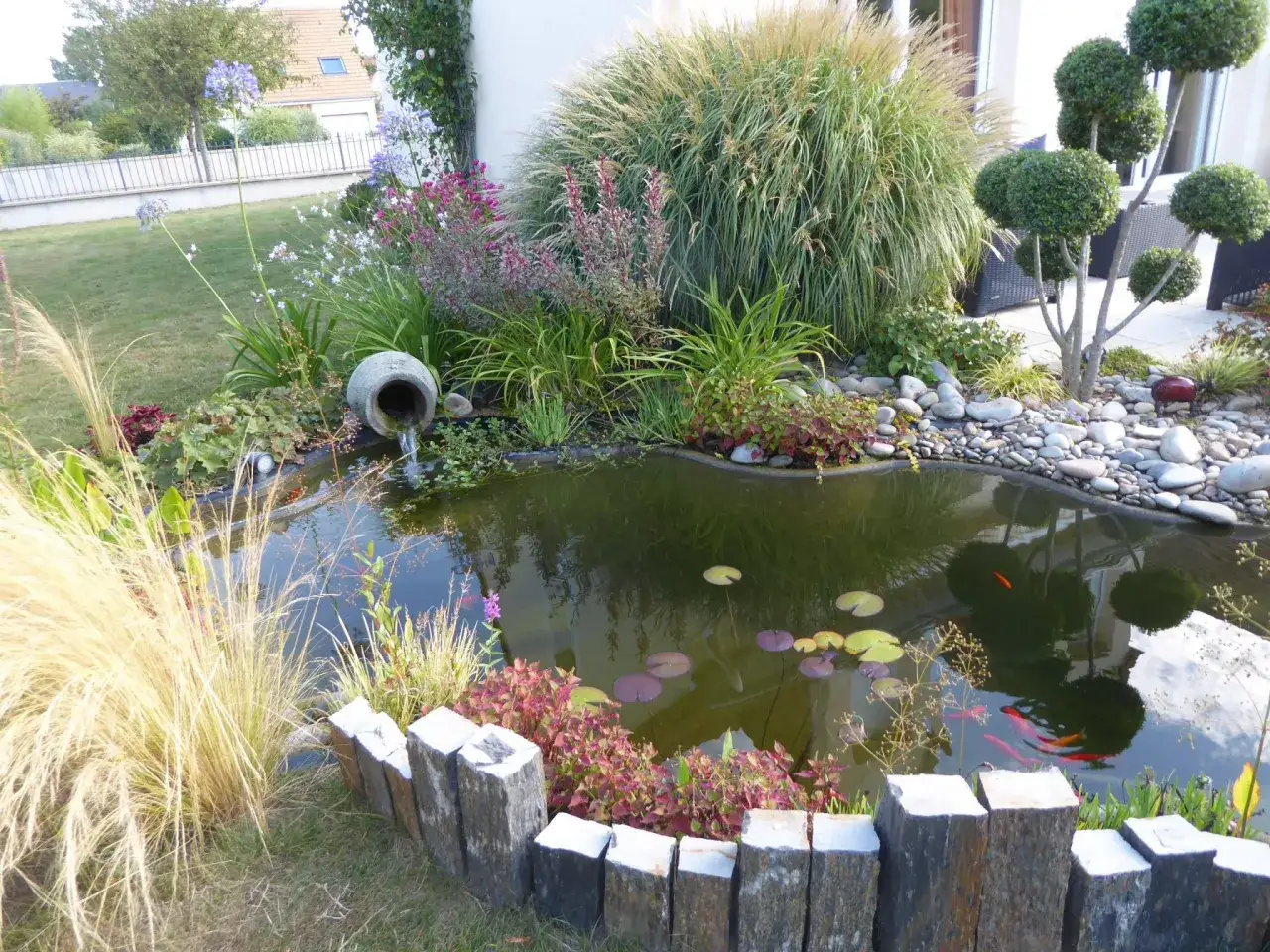 Un petit étang maison avec des poissons rouges, des nénuphars et une cascade en pot. Des herbes hautes et des fleurs entourent ce coin paisible.