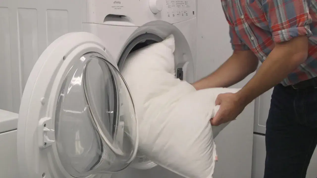 Hombre mete una almohada blanca en una lavadora. Aprende como lavar almohadas para mantenerlas frescas.