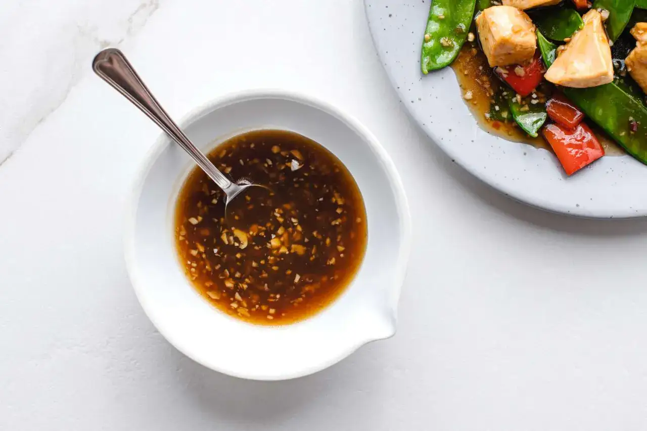 Miseczka z aromatycznym, brązowym sosem chińskim, w którym pływają drobne kawałki czosnku i orzechów. Obok talerz z daniem.
