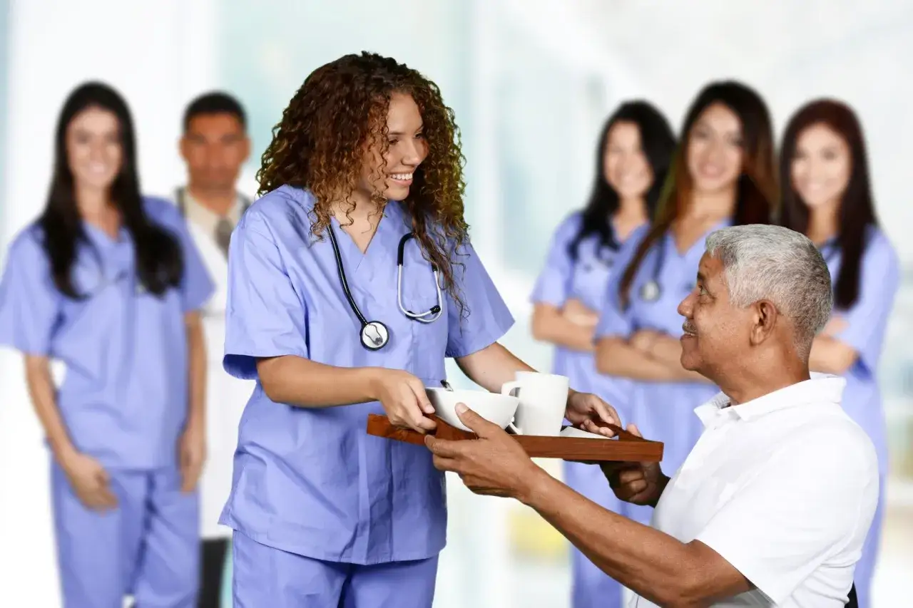 Une jeune femme en formation param&eacute;dicale sans bac, souriante, sert un plateau repas &agrave; un patient. D'autres soignants sont en arri&egrave;re-plan.