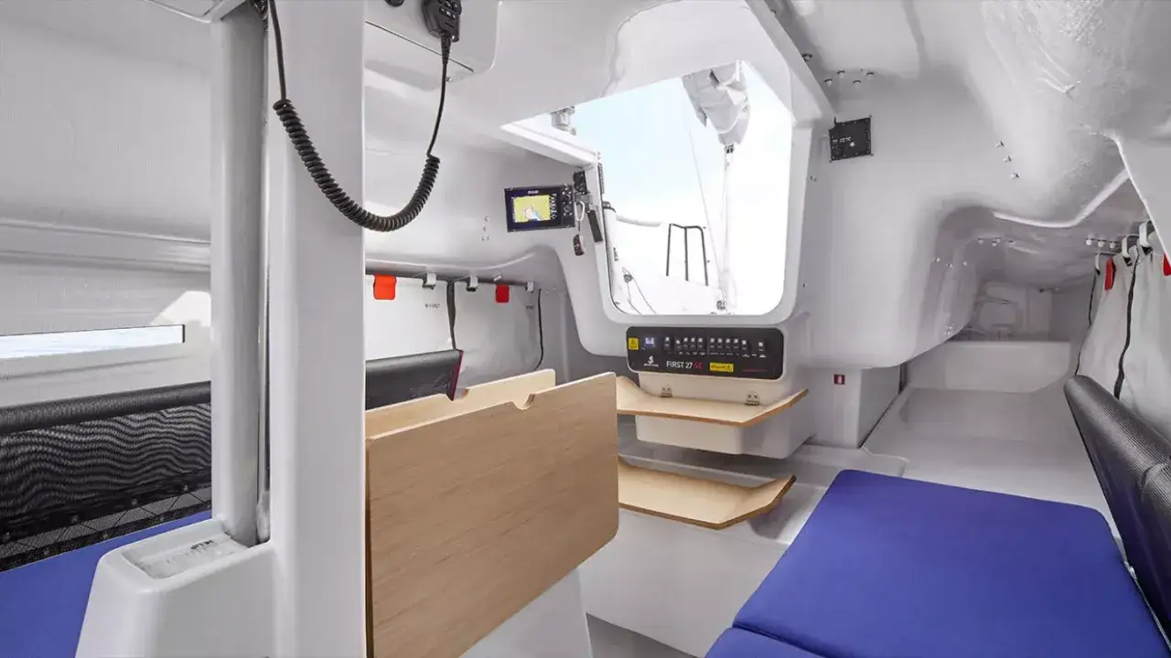 Intérieur du First 27 SE, avec ses banquettes bleues, sa table en bois et son équipement de navigation.