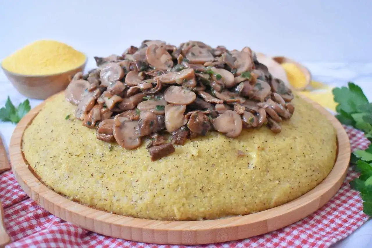 Puszysta polenta z aromatycznymi grzybami duszonymi w sosie, podana na drewnianej desce. Obok miseczka z mąką kukurydzianą.