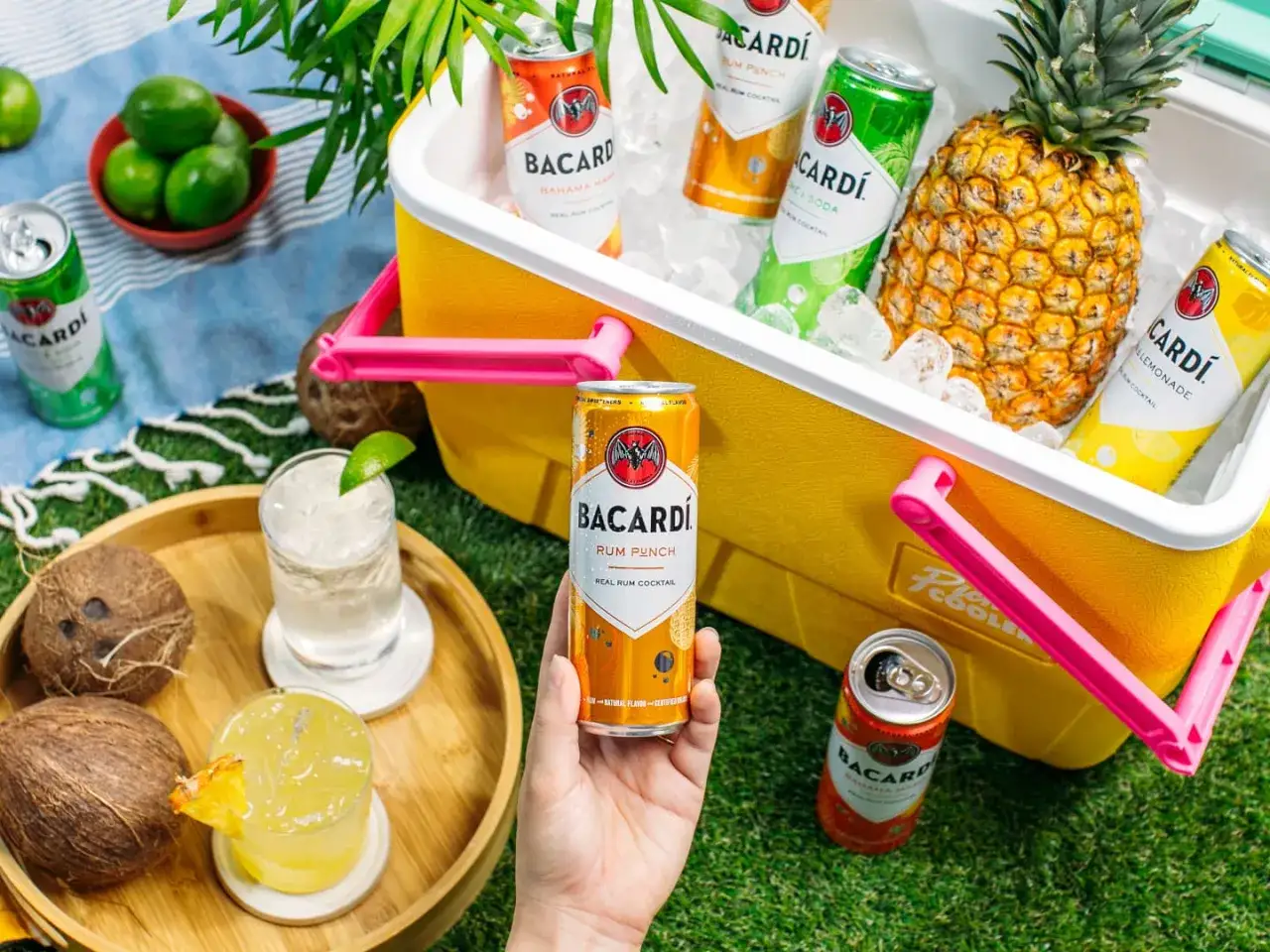 Ręka trzyma puszkę drinka z bacardi Rum Punch. Obok leżą kokosy, drinki i lodówka pełna napojów.