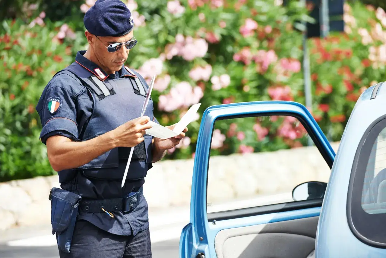 Agente di polizia italiano con giubbotto antiproiettile e berretto blu, legge un documento vicino a un'auto blu. Potrebbe essere un controllo ai sensi dell'art. 180 cds.
