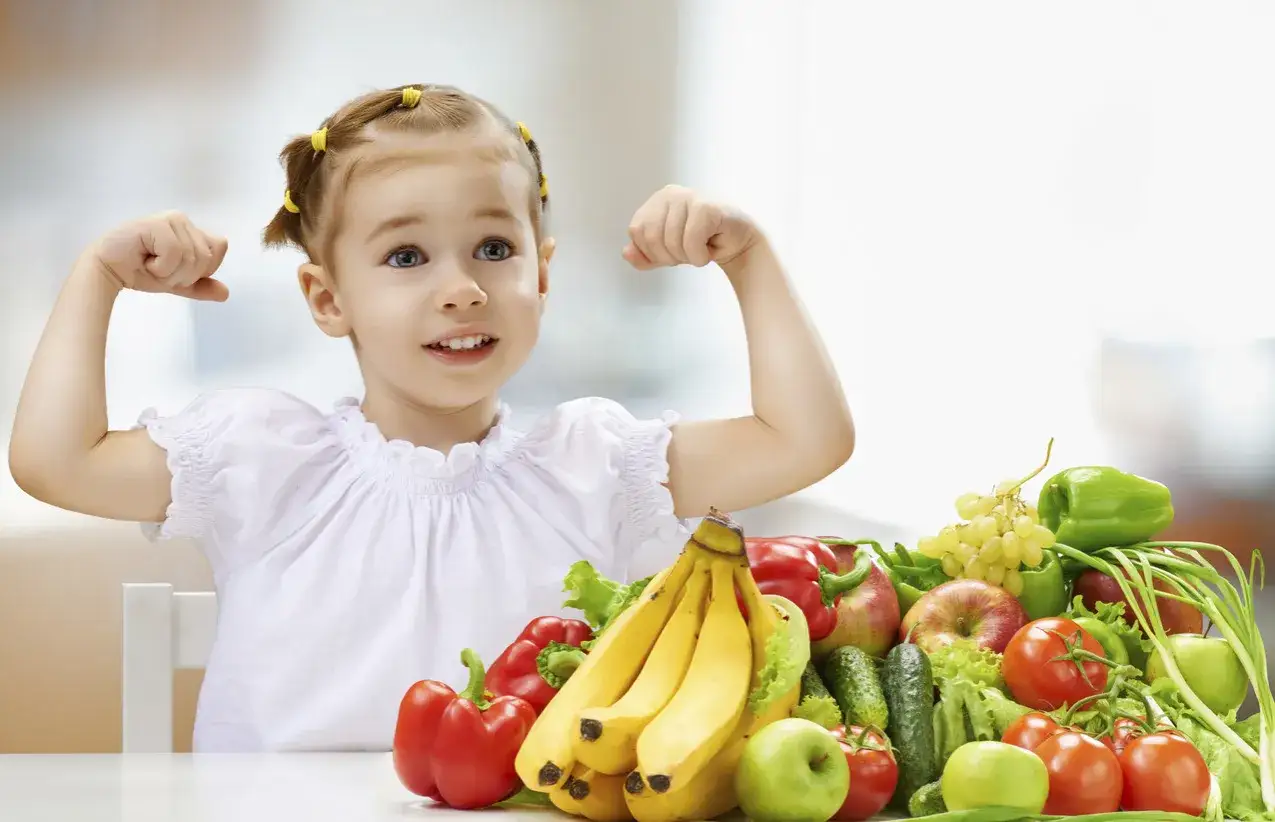 Niña fuerte mostrando sus músculos junto a una variedad de grupos de alimentos para niños: frutas y verduras.