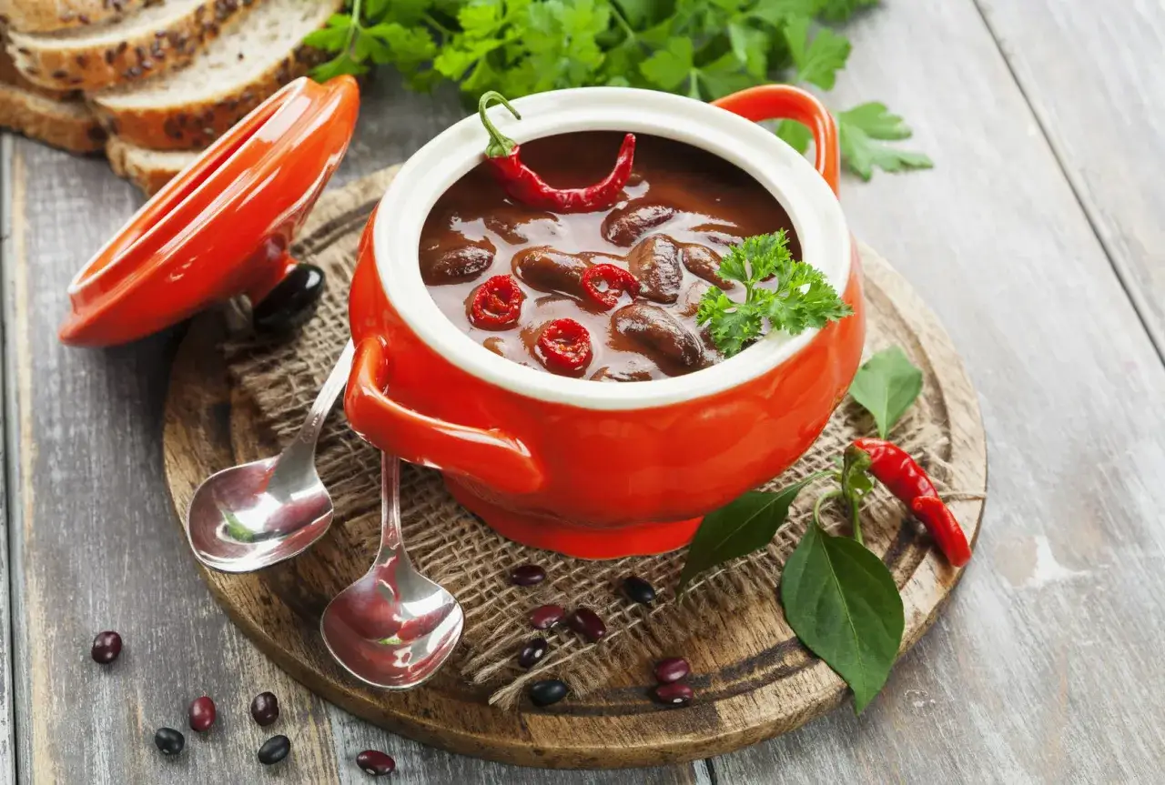 Pyszne chili con carne w pomarańczowej miseczce, ozdobione papryczką chili i natką pietruszki. Obok kromki chleba i łyżeczki.