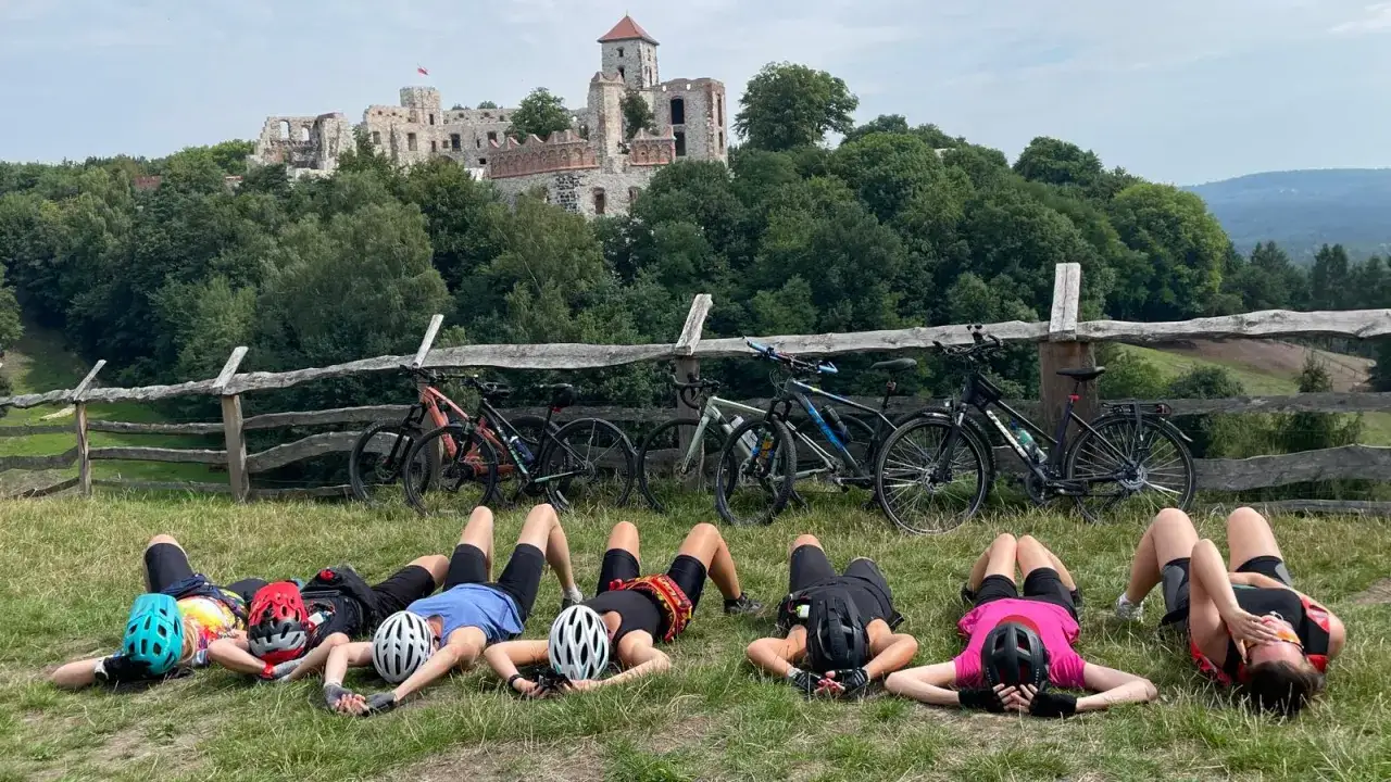 Odpoczynek po rowerowej przygodzie na trasie rowerowej Szlak Orlich Gniazd. Grupa rowerzystów leży na trawie przed ruinami zamku.