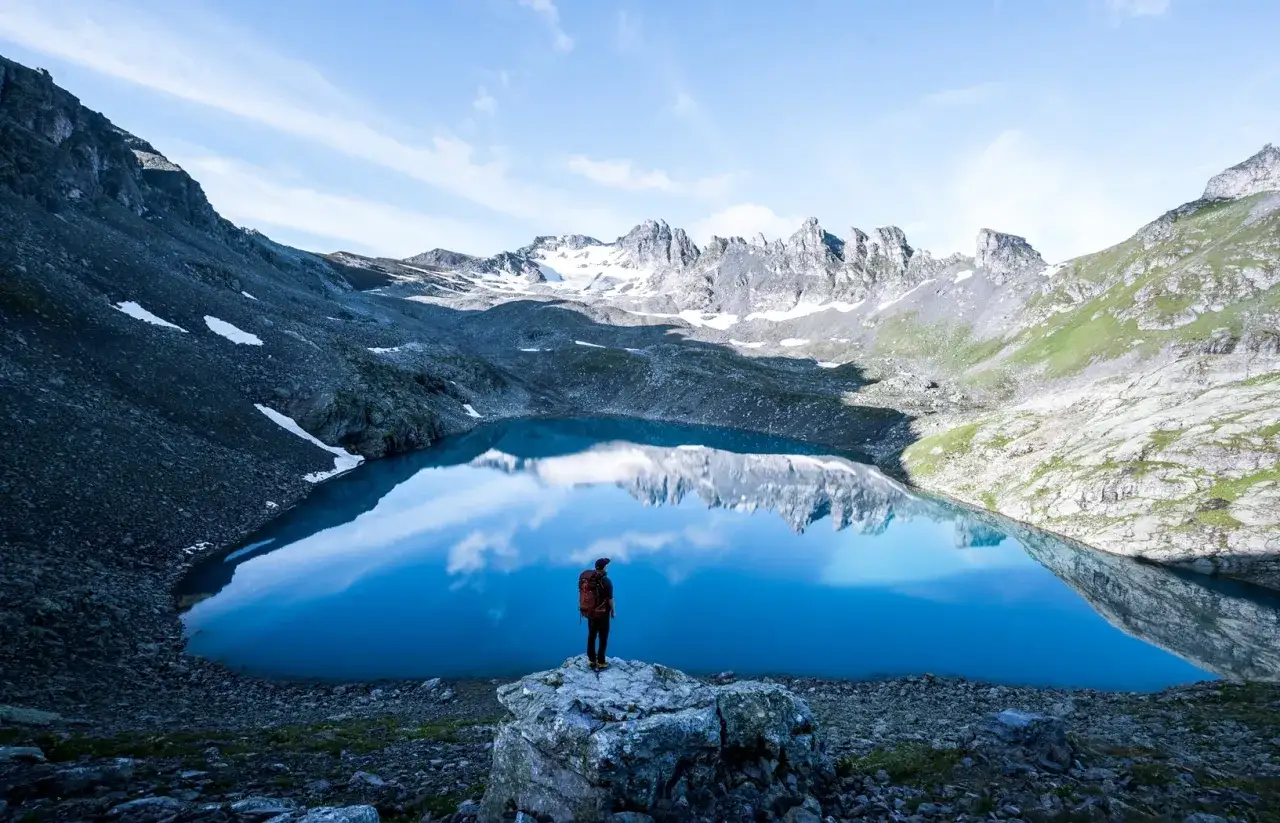 Ein Wanderer steht am Ufer eines spiegelglatten Bergsees in den Schweizer Alpen. Die majest&auml;tischen Gipfel spiegeln sich im tiefblauen Wasser.