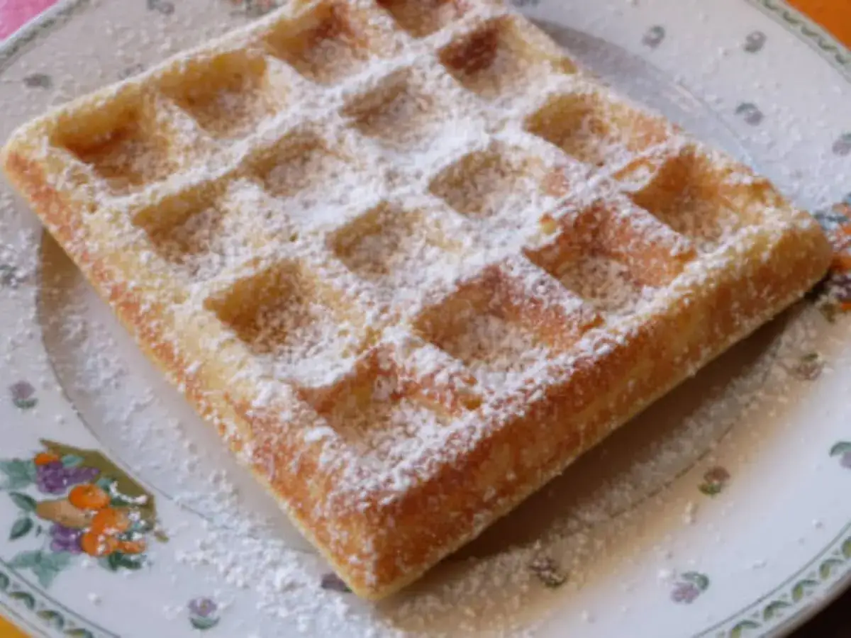 Une gaufre lyonnaise dorée, saupoudrée de sucre glace, repose sur une assiette décorée de fleurs. Un délice simple et gourmand.