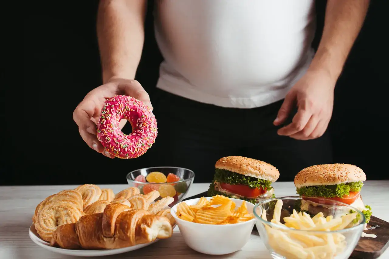 Ręka trzymająca różowego pączka nad stołem pełnym słodyczy i fast foodów. Obraz symbolizuje kompulsywne objadanie się.