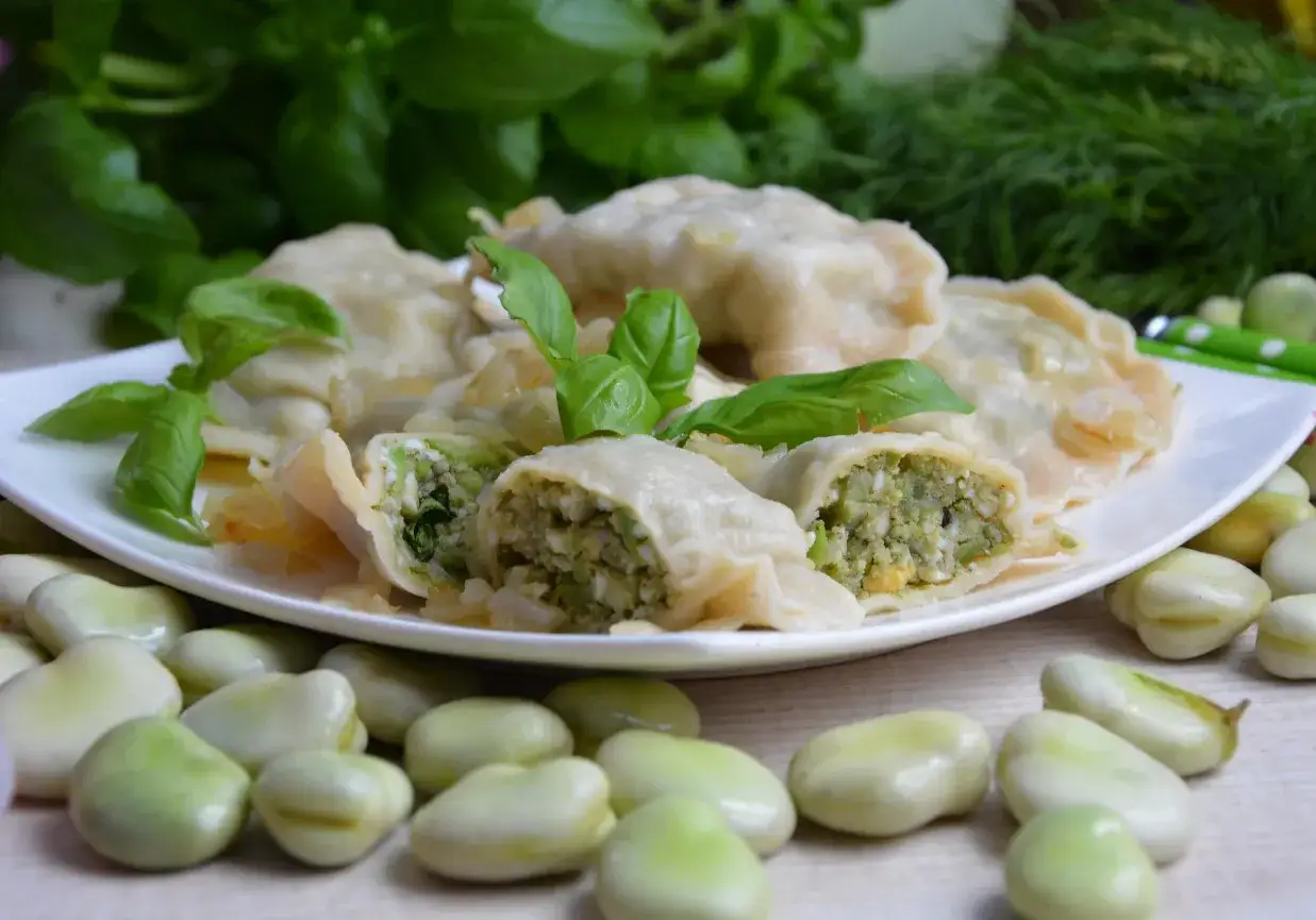 Pyszne pierogi z bobem, jajkiem i ziołami, udekorowane świeżą bazylią. Na talerzu widać też kilka fasolek bobu.