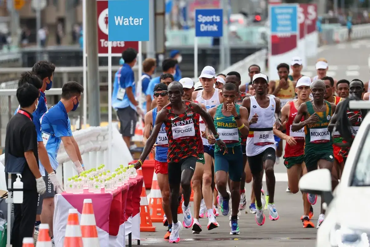 Biegacze na trasie maratonu, Eliud Kipchoge z Kenii biegnie po wodę. Czyżby przed bieganiem wziął aspirynę dla lepszej wydajności?