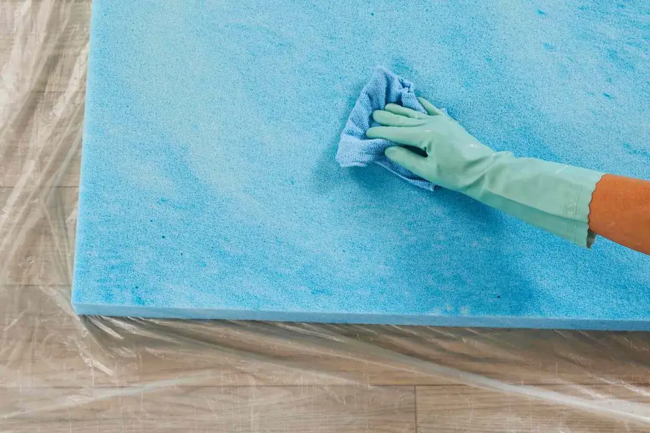 Main bleue, gant vert, chiffon bleu nettoient un matelas en mousse. Apprenez comment nettoyer un matelas en mousse pour une fra&icirc;cheur retrouv&eacute;e.
