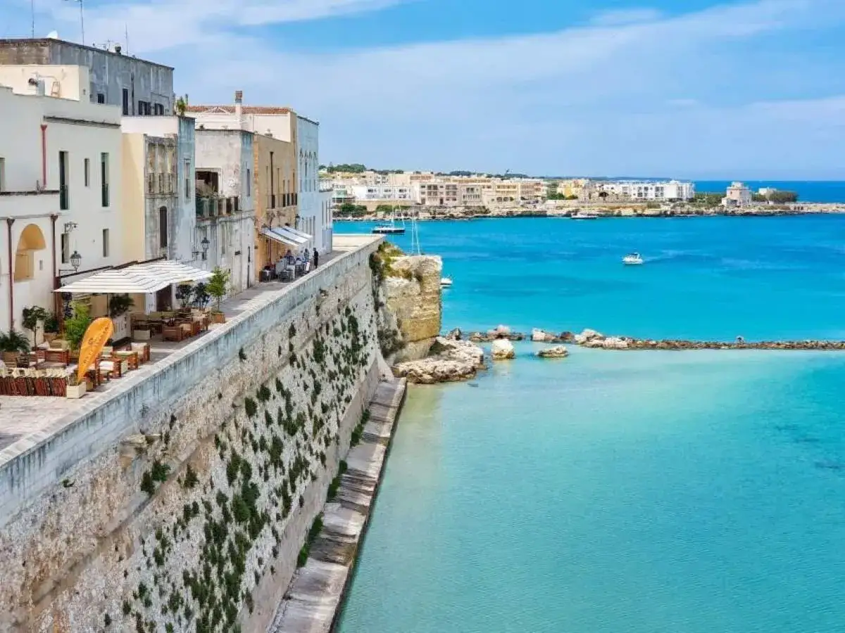 Vista del lungomare di Otranto centro, con edifici bianchi affacciati sul mare turchese e un ristorante all'aperto.