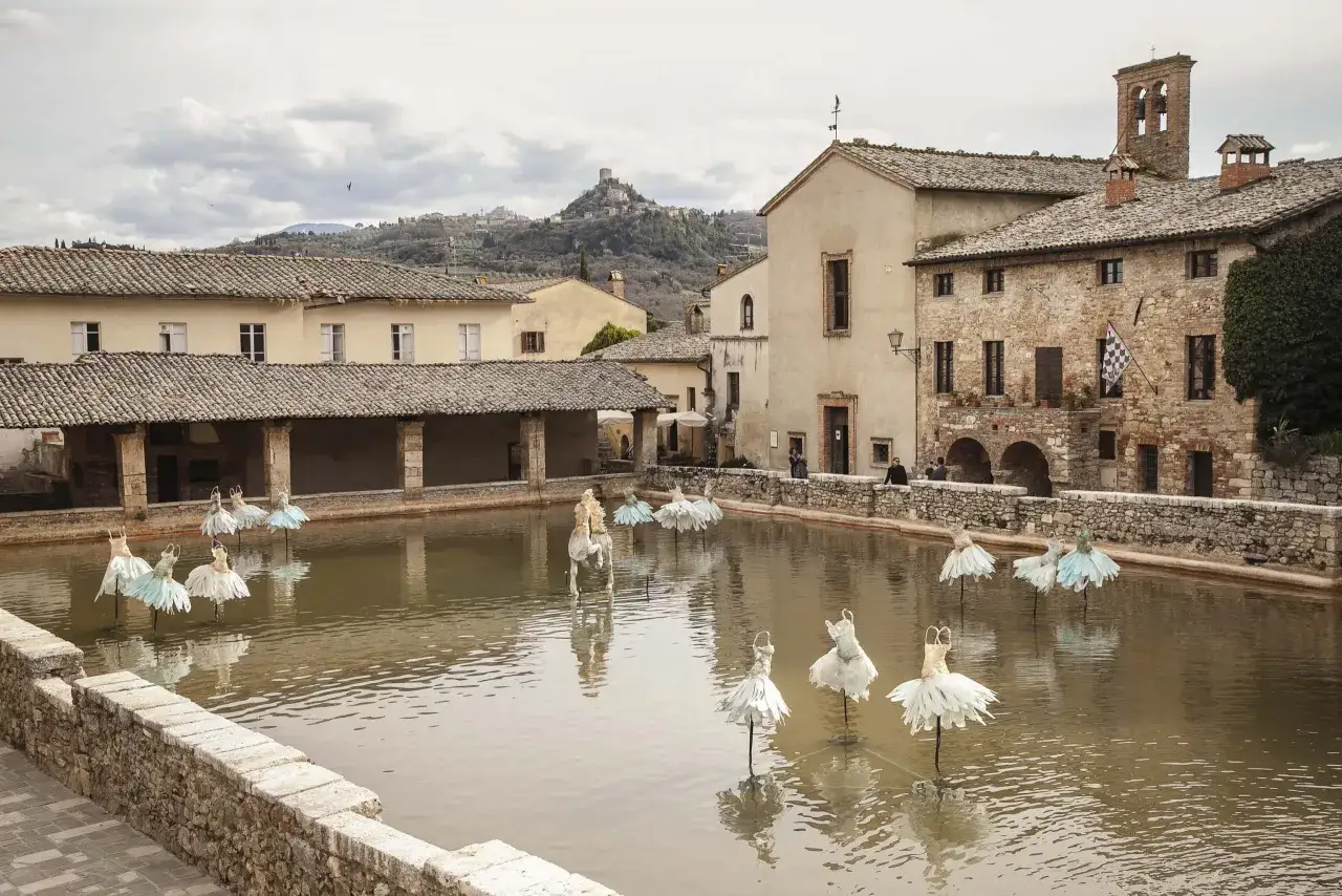 Bagno Vignoni: cosa vedere? La vasca termale con installazioni artistiche di abiti da ballerina e un cavallo bianco, sullo sfondo il borgo toscano.