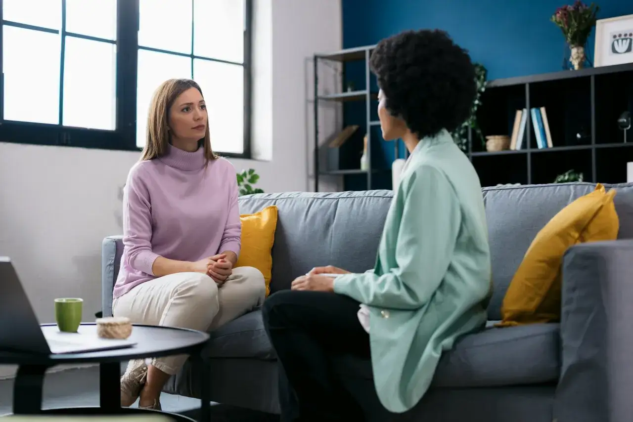 Dos mujeres conversan en un sof&aacute;. Una tiene el pelo casta&ntilde;o y lleva un jersey lila. La otra tiene el pelo afro y viste una chaqueta verde menta. Son **im&aacute;genes para describir** una sesi&oacute;n de terapia.