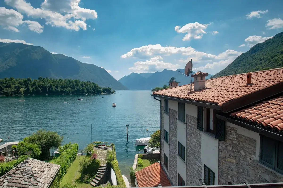 Vue d'une chambre d'h&ocirc;te en Lombardie, avec un lac paisible, des montagnes verdoyantes et un ciel bleu parsem&eacute; de nuages.