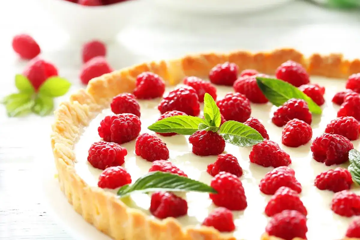 Tarta z kremem z białej czekolady, ozdobiona malinami i listkami mięty, wygląda apetycznie.