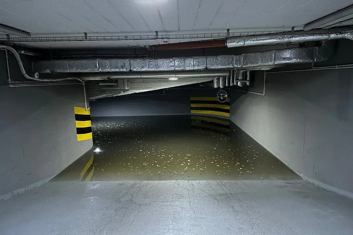 Podziemny parking zalany wodą po powodzi błyskawicznej. Widać żółto-czarne słupki ostrzegawcze i instalacje wentylacyjne pod sufitem.