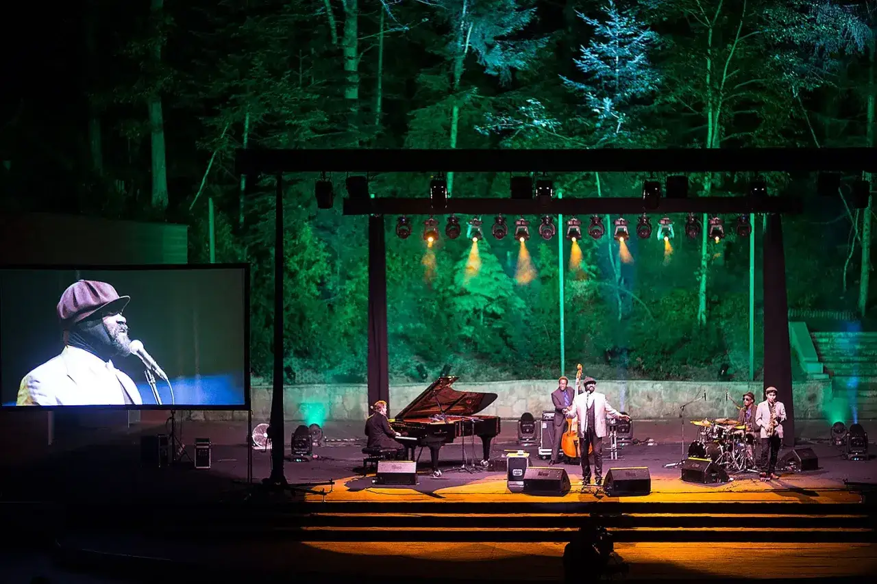 Gregory Porter koncert w otoczeniu drzew. Na scenie muzycy, w tym wokalista, pianista, basista i perkusista. Na ekranie widać zbliżenie na Gregory'ego Portera.