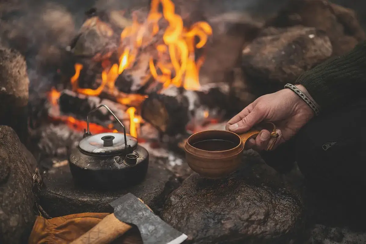 Vid lägerelden värms en kaffepanna, redo för att brygga kokkaffe. En träkopp med varm dryck hålls i handen.