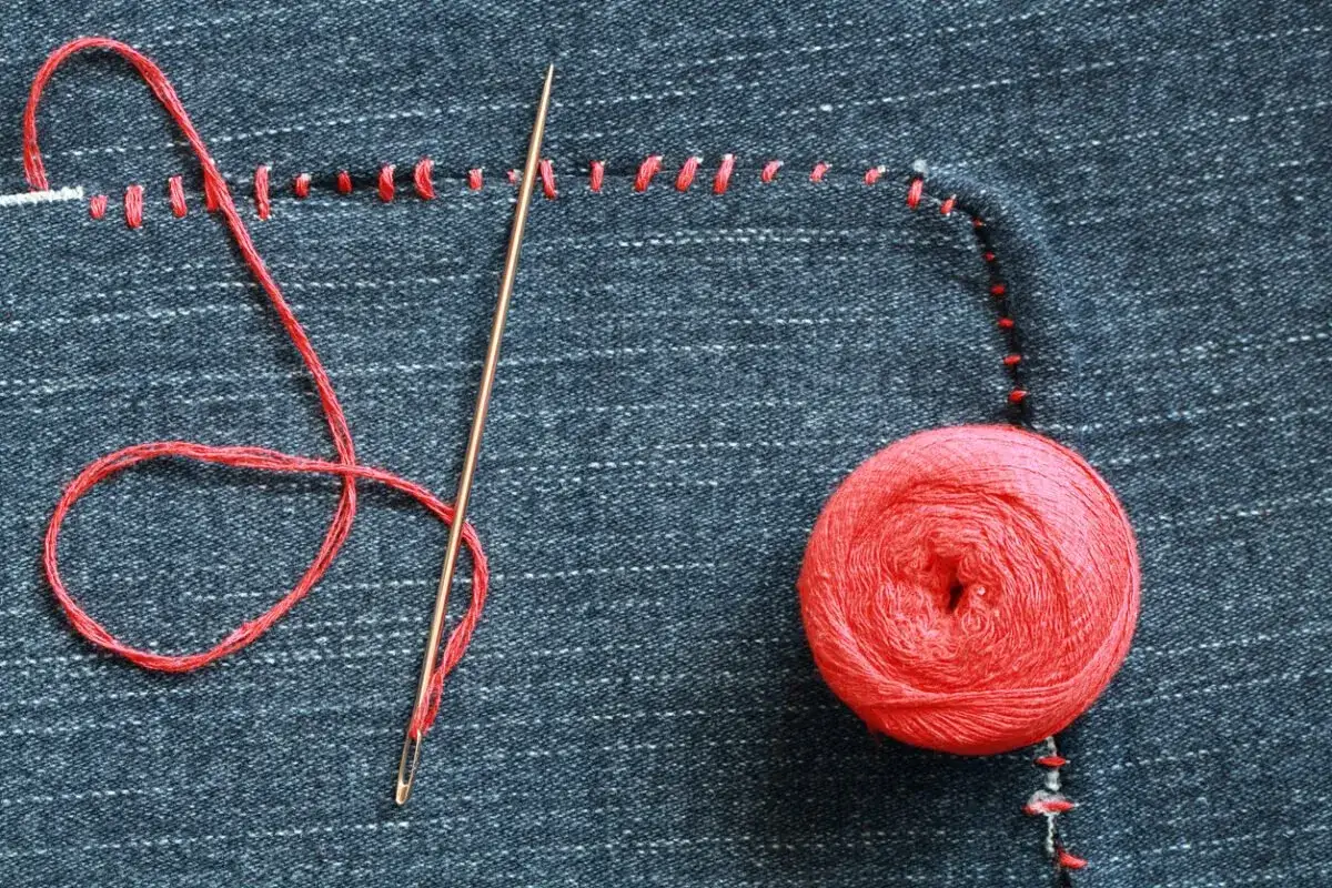 Hilo rojo, aguja y ovillo sobre tela vaquera, mostrando cómo coser un agujero a mano con puntadas visibles.