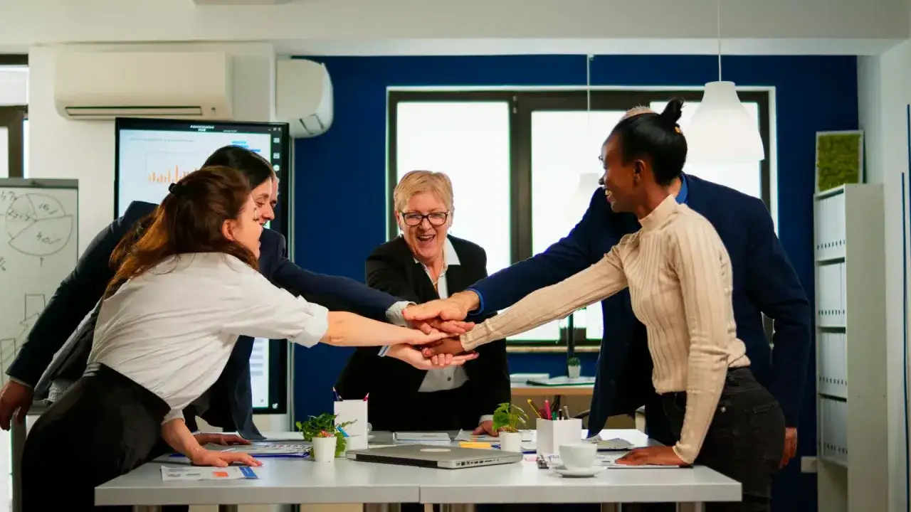 A diverse team in a modern office stacks hands, showing unity. The supervisor, smiling, leads this gesture, embodying the role of fostering collaboration and shared success.