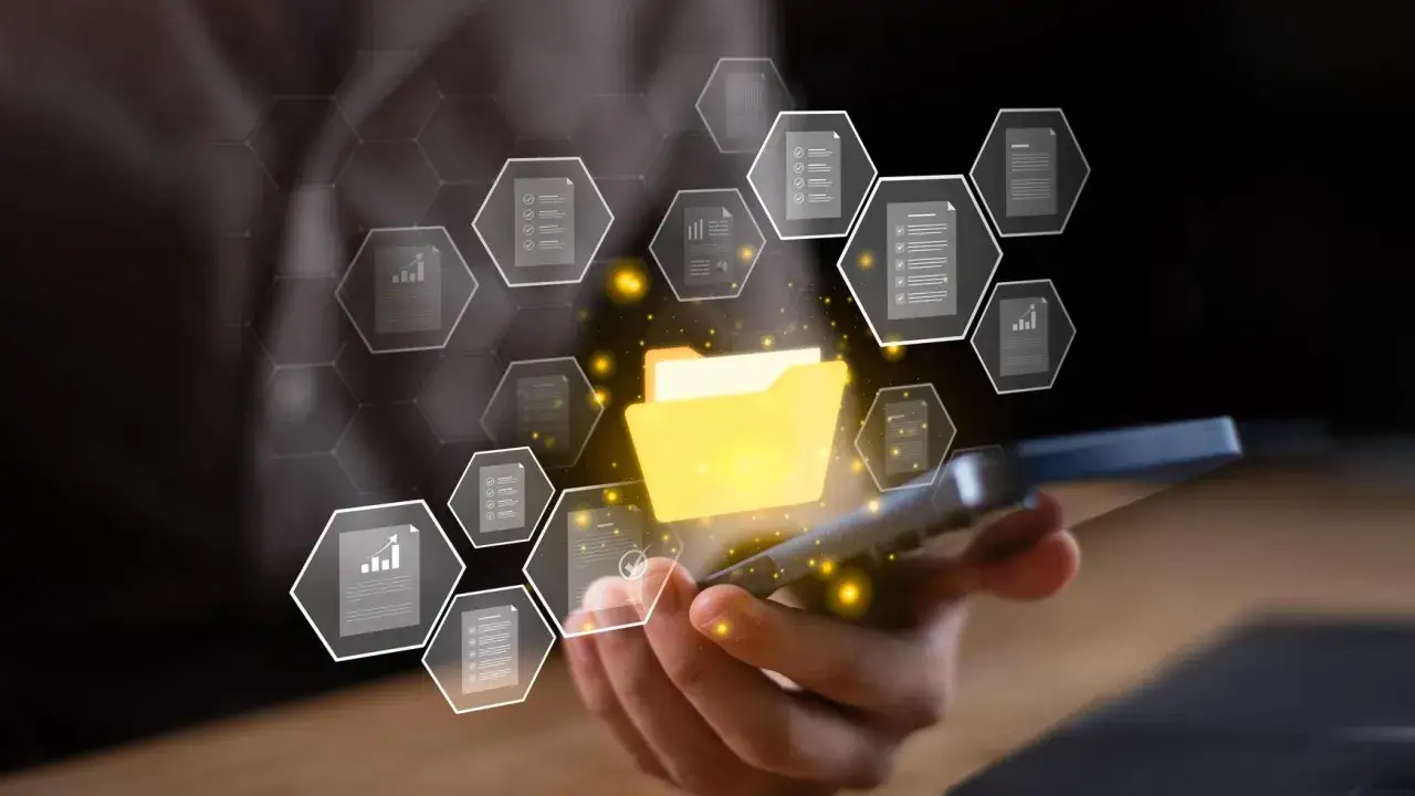 Dłoń trzyma smartfon, z którego wyrasta świecący folder z dokumentami. Wokół unoszą się ikony plików, tworząc cyfrową sieć. To wizualizacja zdjęć w chmurze.
