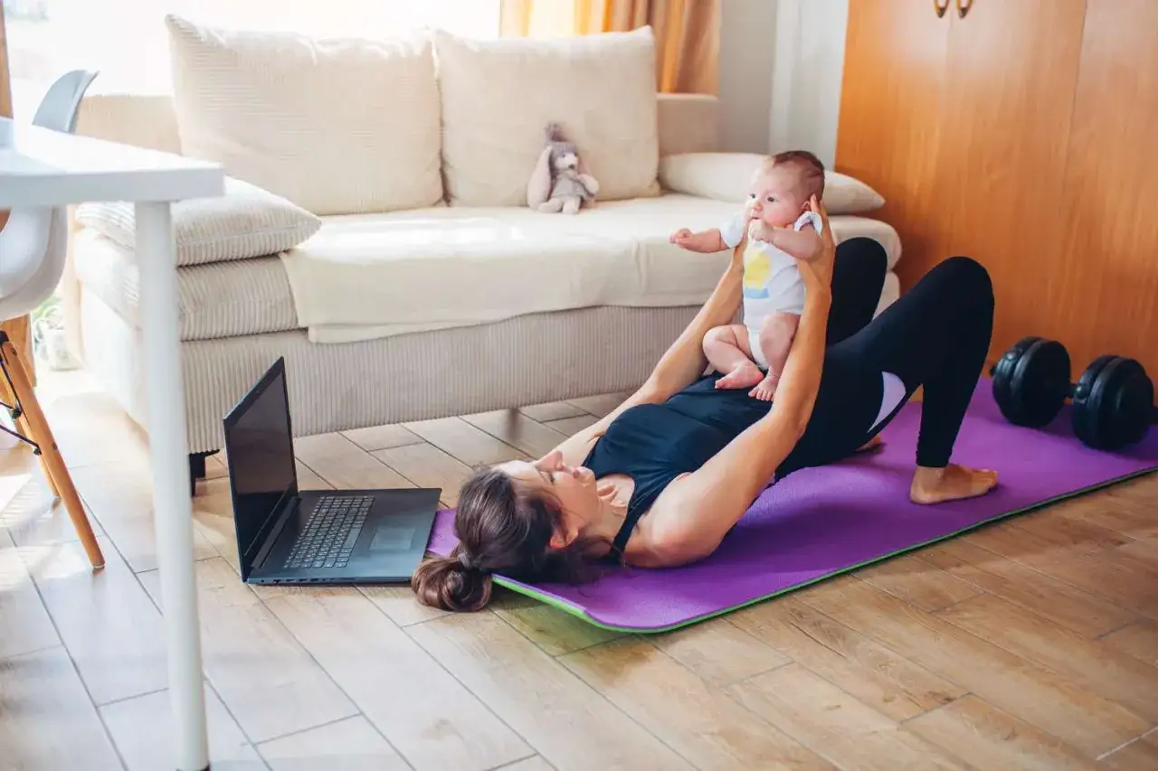 Mamma tr&auml;nar hemma med bebis, en del av hennes resa att g&aring; ner i vikt efter graviditet. Tr&auml;ningsmatta, laptop och hantlar syns.
