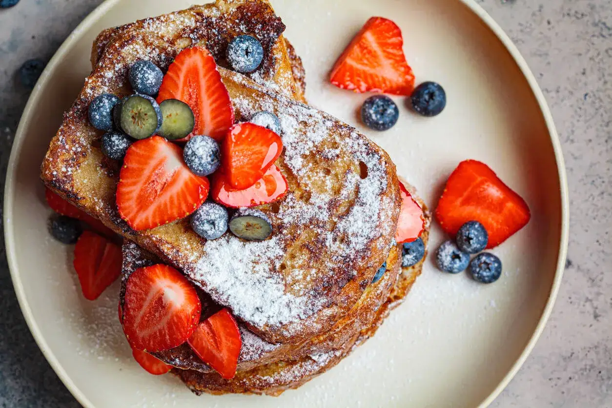 En stapel gyllene fattiga riddare, toppad med f&auml;rska jordgubbar och bl&aring;b&auml;r, pudrad med florsocker. Ett perfekt recept p&aring; french toast f&ouml;r en h&auml;rlig frukost.