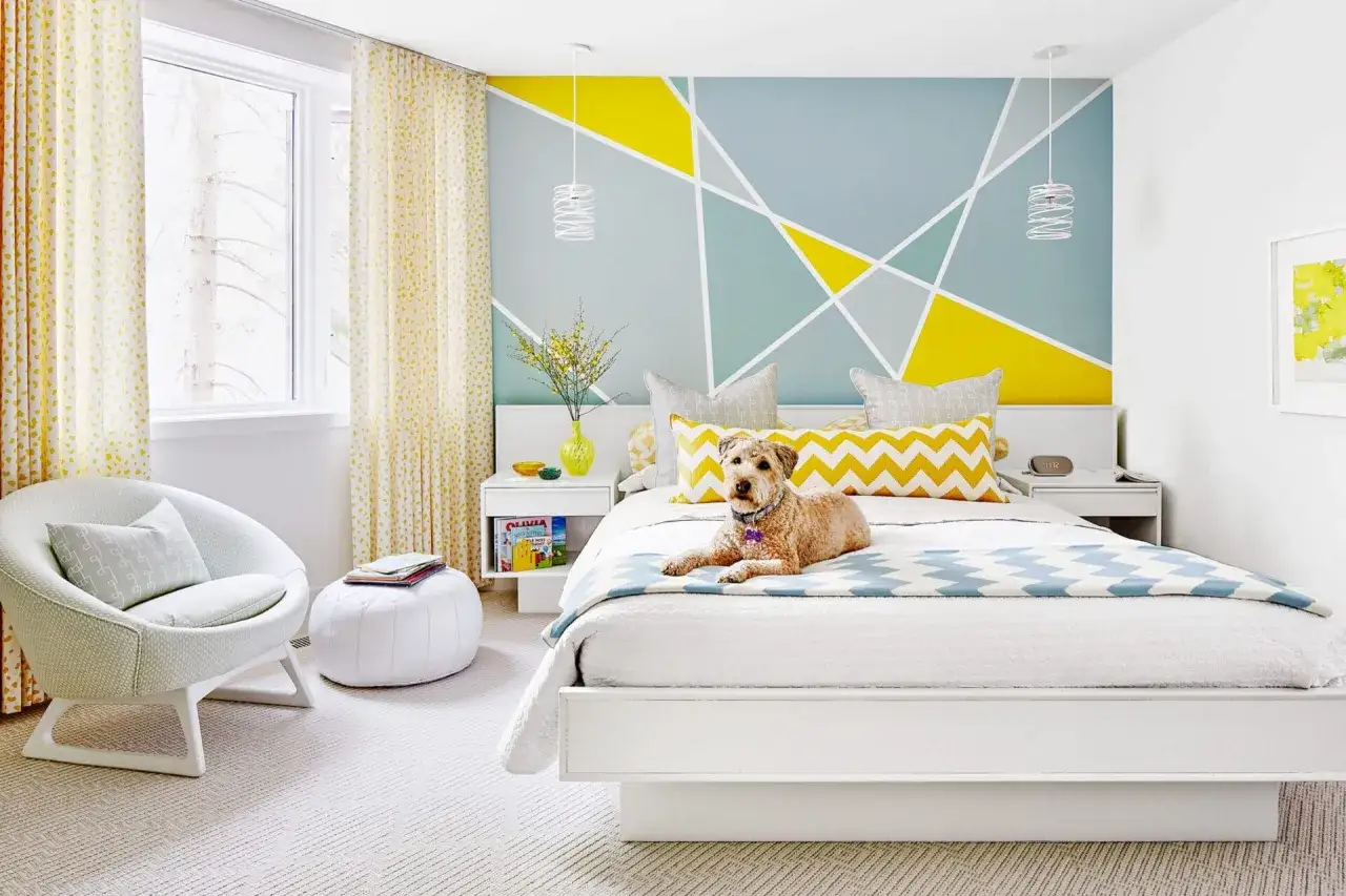 Un chien sur un lit dans une chambre lumineuse. Le mur derri&egrave;re le lit pr&eacute;sente une id&eacute;e motif peinture murale g&eacute;om&eacute;trique jaune et bleu.
