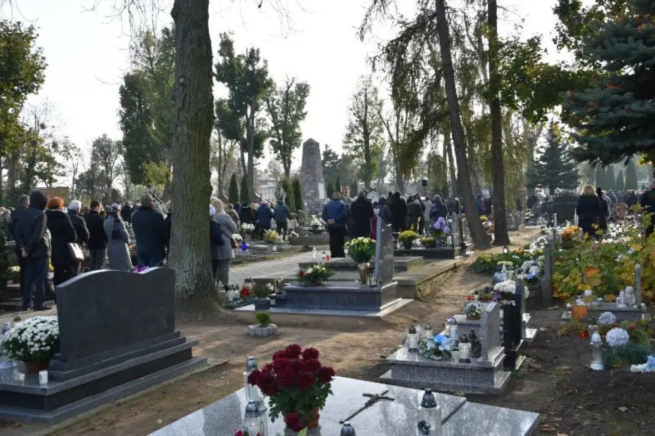 Uroczystość na cmentarzu parafialnym, gdzie zgromadzeni ludzie oddają hołd bliskim. W tle widać nagrobki i drzewa.