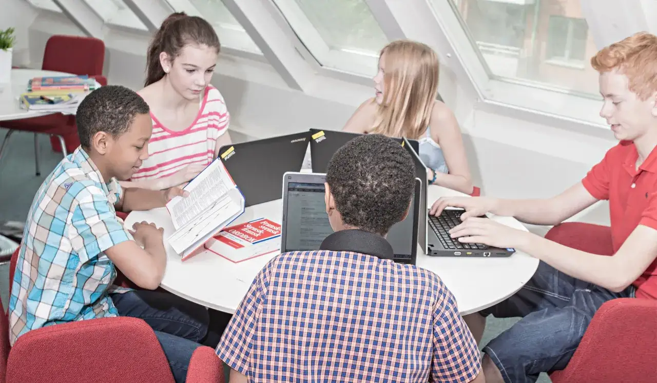 Barn samarbetar kring en **academy online**-uppgift med b&ouml;cker och laptops.