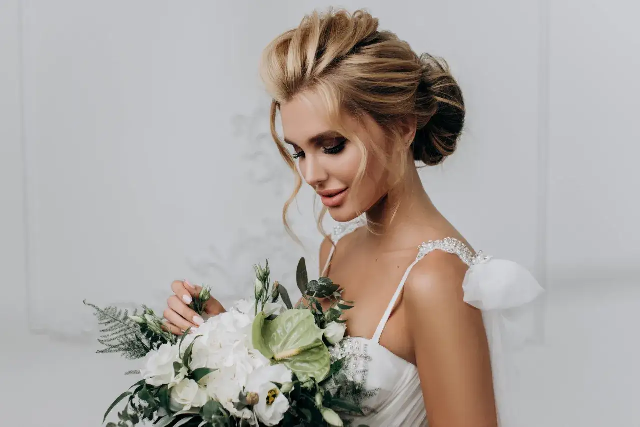 Elegancka panna młoda z bukietem kwiatów, prezentująca proste fryzury ślubne. Jej blond włosy są upięte w stylowy kok, a delikatne loki opadają na ramiona.