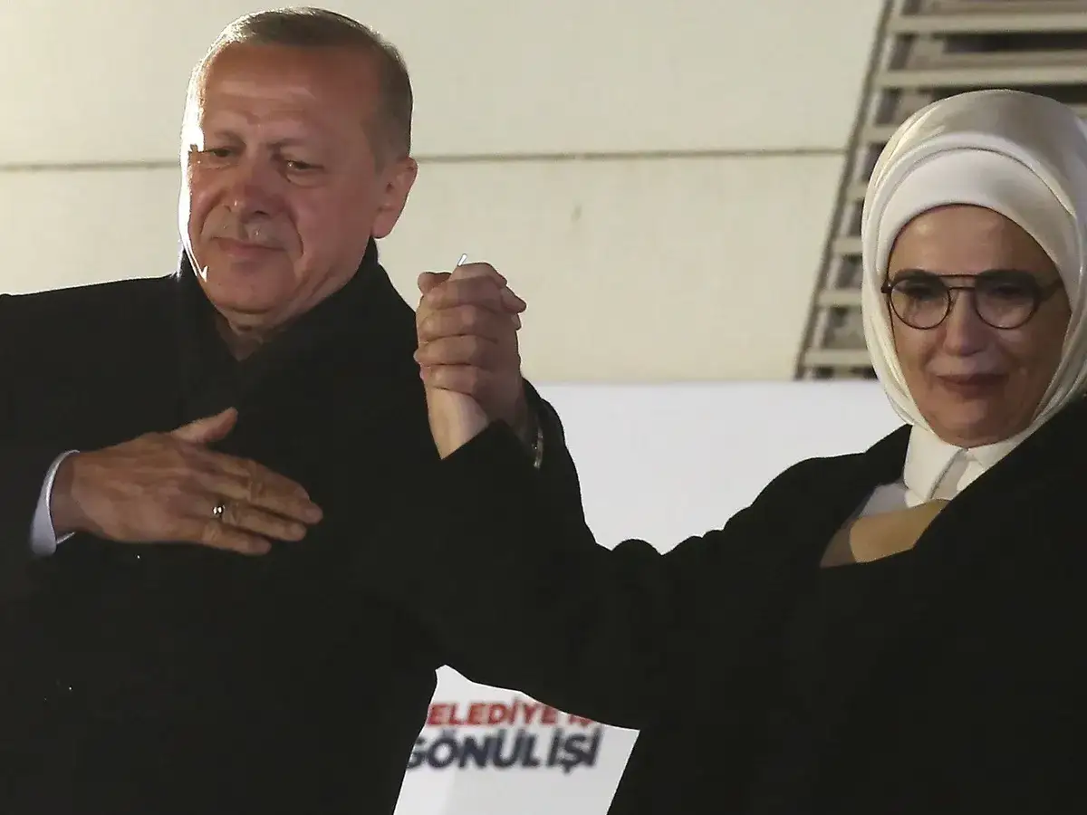 Erdogan hält die Hand seiner Frau, die ein Kopftuch trägt. Sie lächeln und scheinen sich zu freuen.