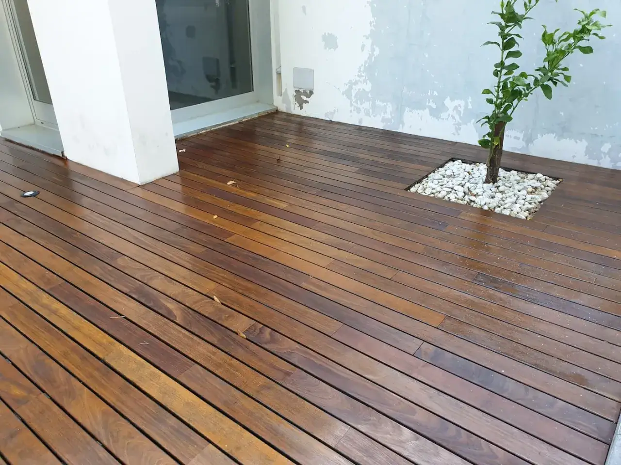 Elegante terraza de maderas para exterior, con un &aacute;rbol joven rodeado de piedras blancas.