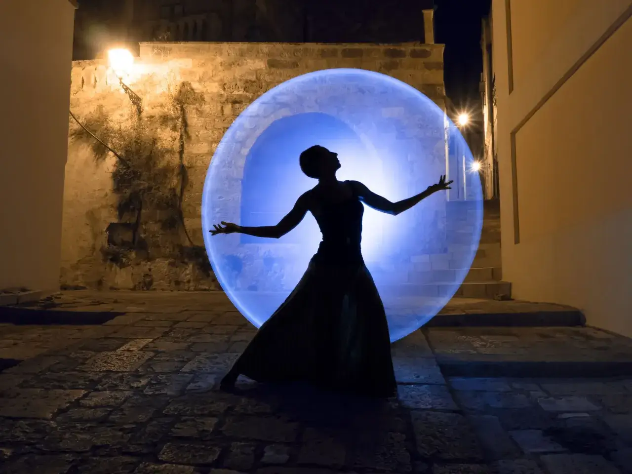 Une silhouette de danseuse dans une bulle de lumière bleue, une photo light painting dans une ruelle pavée.