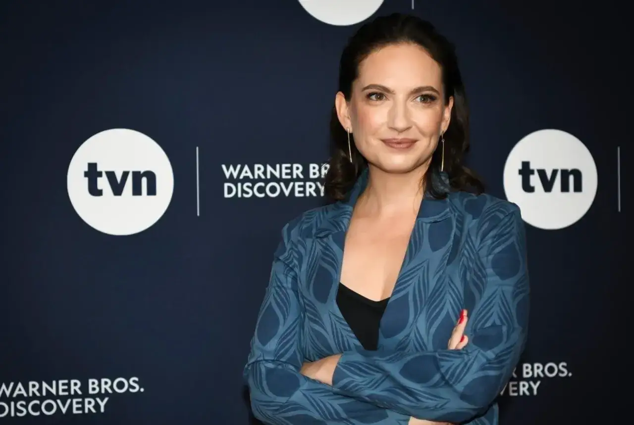 Anna Cieślak w niebieskiej marynarce z piórami, z uśmiechem pozuje na tle logo TVN i Warner Bros. Discovery.
